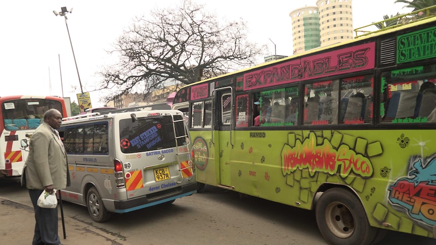 Man waits on footpath by green, loud bus