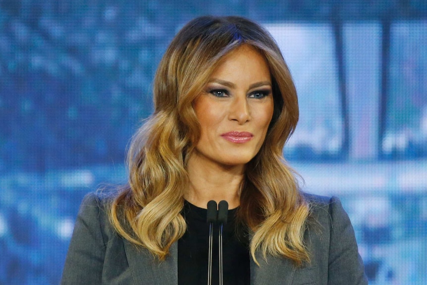 Melania Trump stands in front of a microphone in front of a blue background.