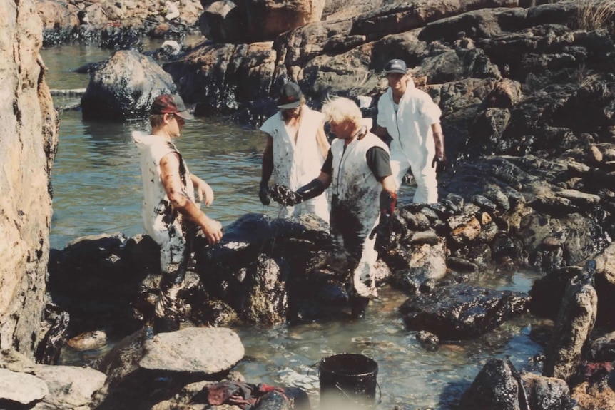 Four people wearing white jumpsuits cleaning up an oil spill on a beach.
