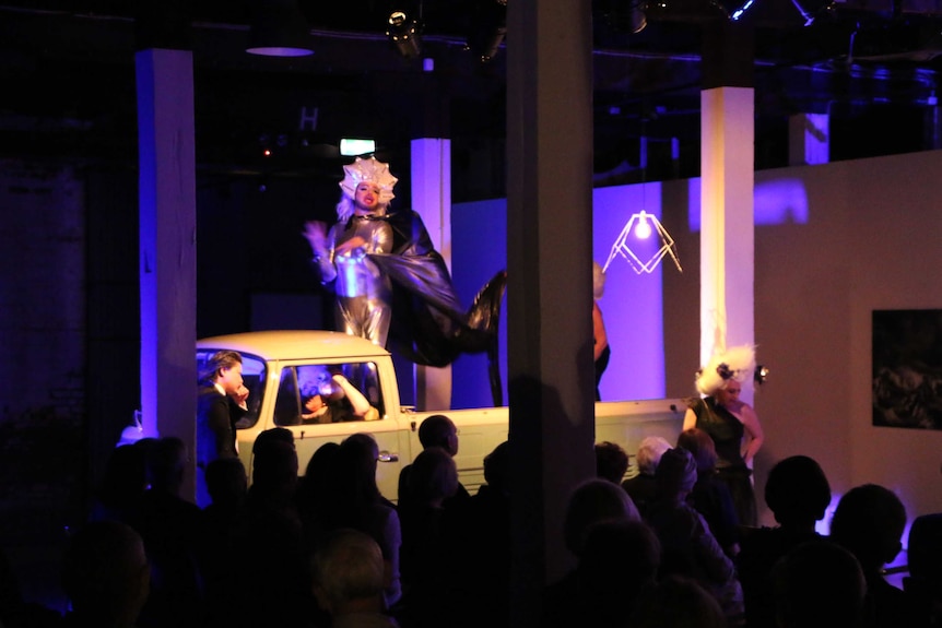 A man dressed in a Priscilla costume stands on the back of a ute in the theatre.