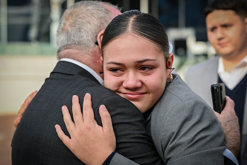 An older man stands with a teenage girl in a school uniform, hugging her and consoling her