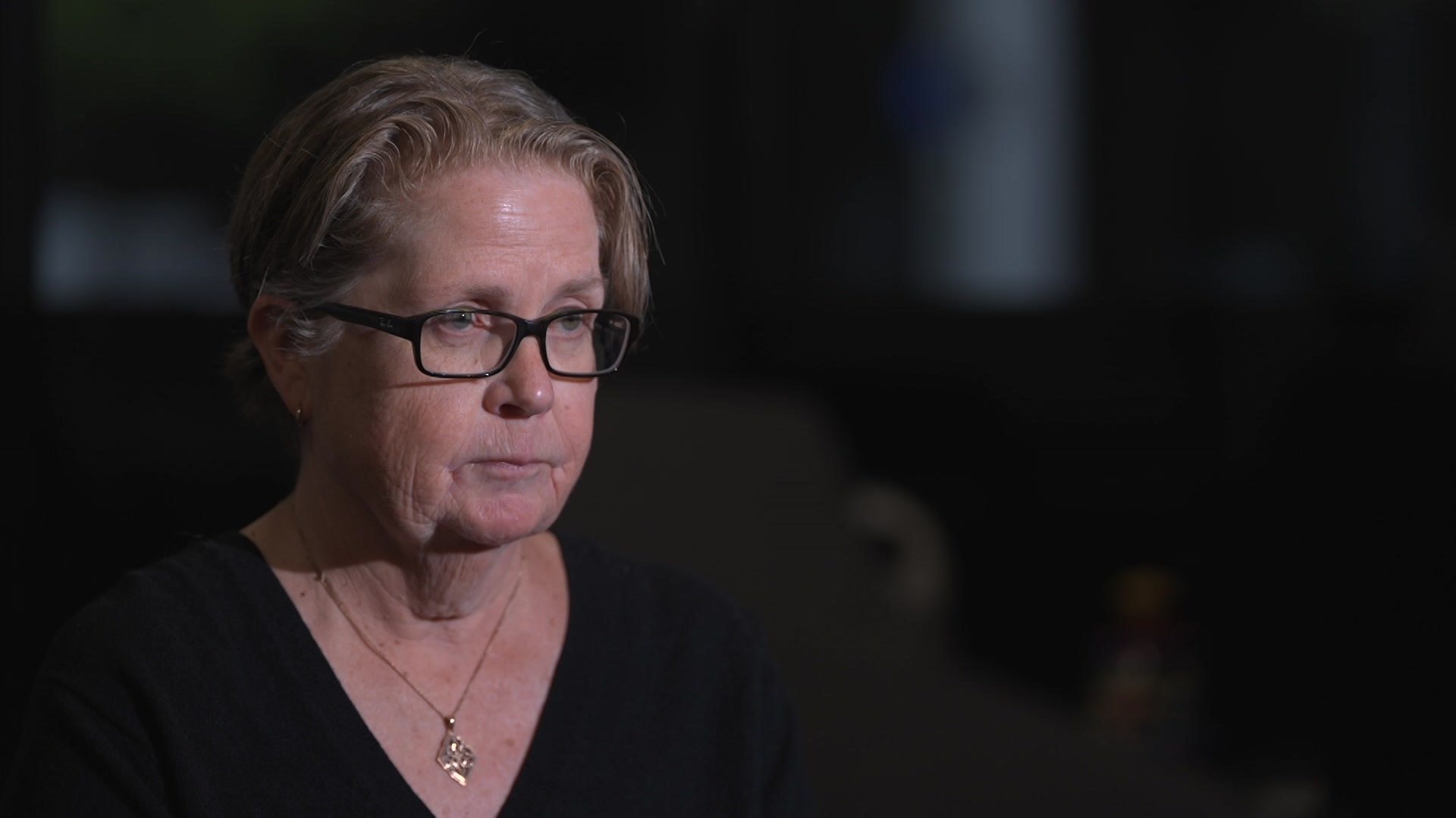 A woman with short blonde hair and black-framed glasses sits in a dark room looking serious.