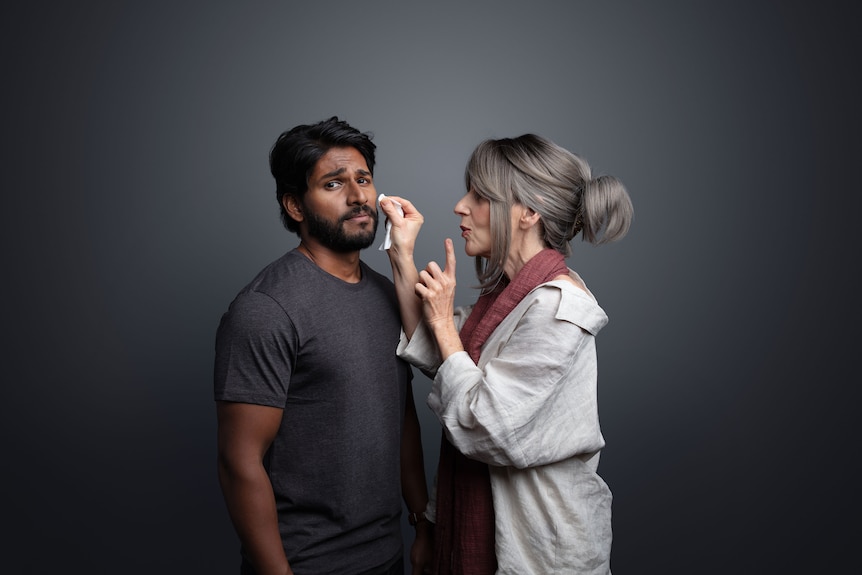 A brown man with dark hair wears a grey t-shirt and winces at the camera while a white woman with grey hair dabs his face.