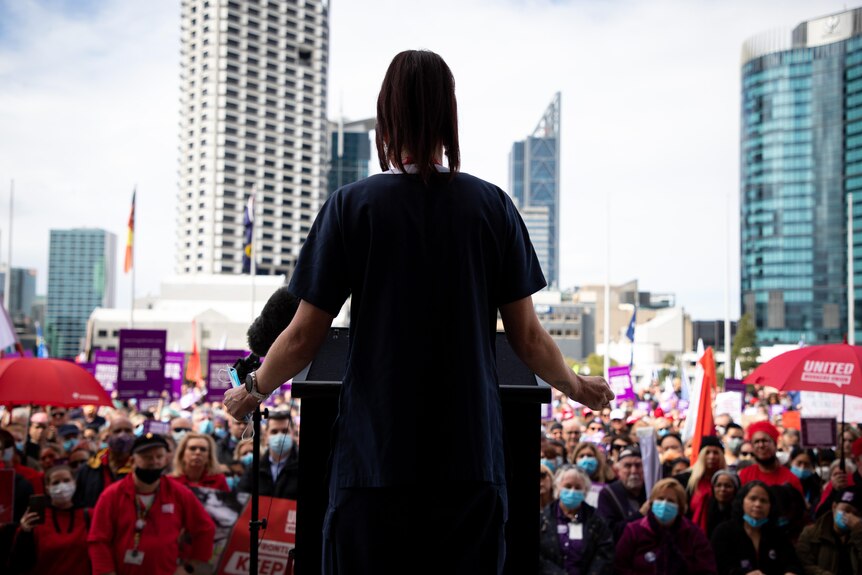 Una enfermera fotografiada de espaldas se dirige a una gran multitud en un mitin