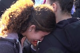 A young woman cries as she rests her head on the shoulder of another woman amongst a crowd of people.