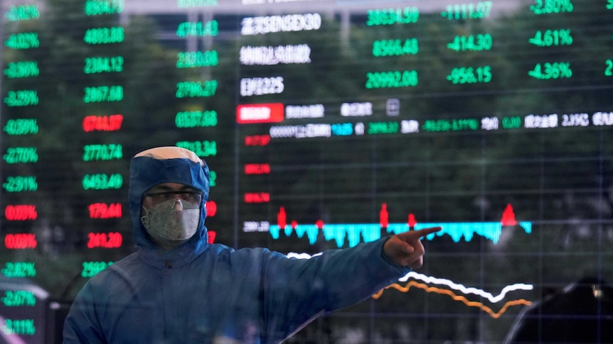 A man in a blue hazmat suit with a facemask and goggles stands in front of a stock exchange board.