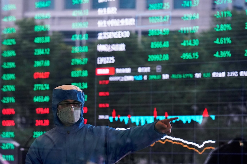 A man in a blue hazmat suit with a facemask and goggles stands in front of a stock exchange board.