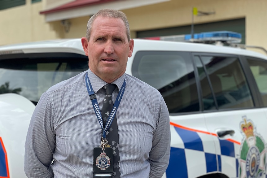 Policeman stands in front of marked police car with serious expression on his face.