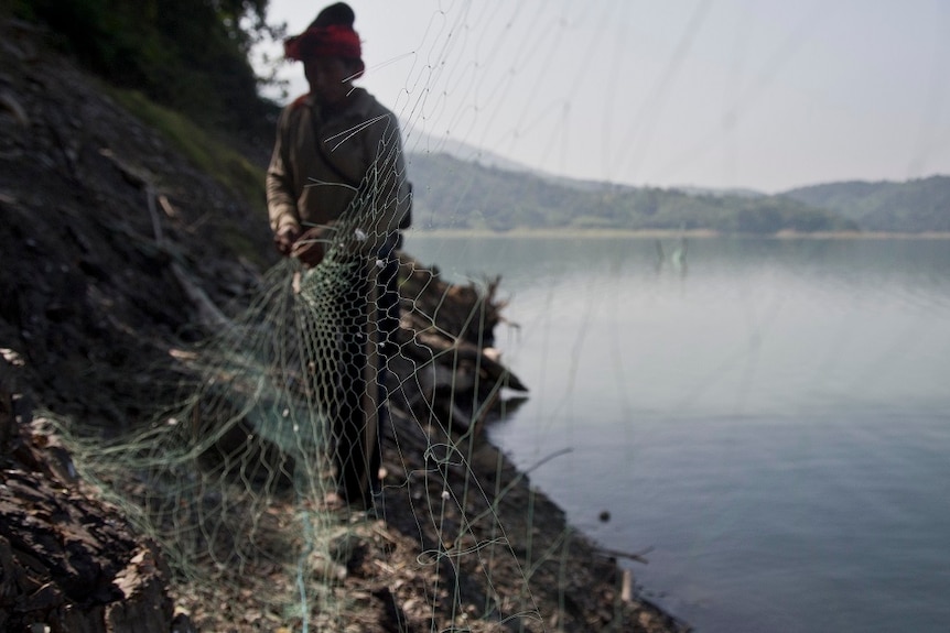 A man unhitches a waist-high net from supporting stakes rigged into the banks of a reservoir.