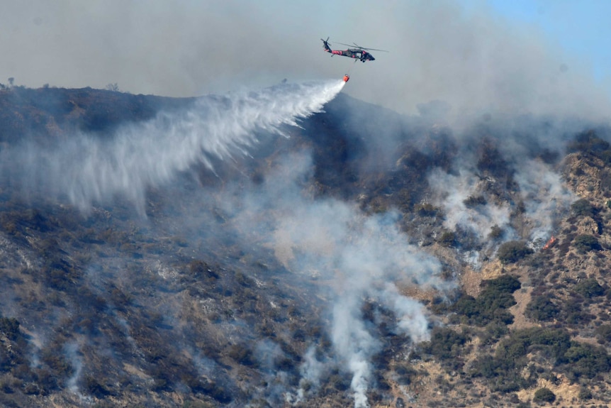A helicopter flies above a burning hill, dropping flame retardant on the blaze.