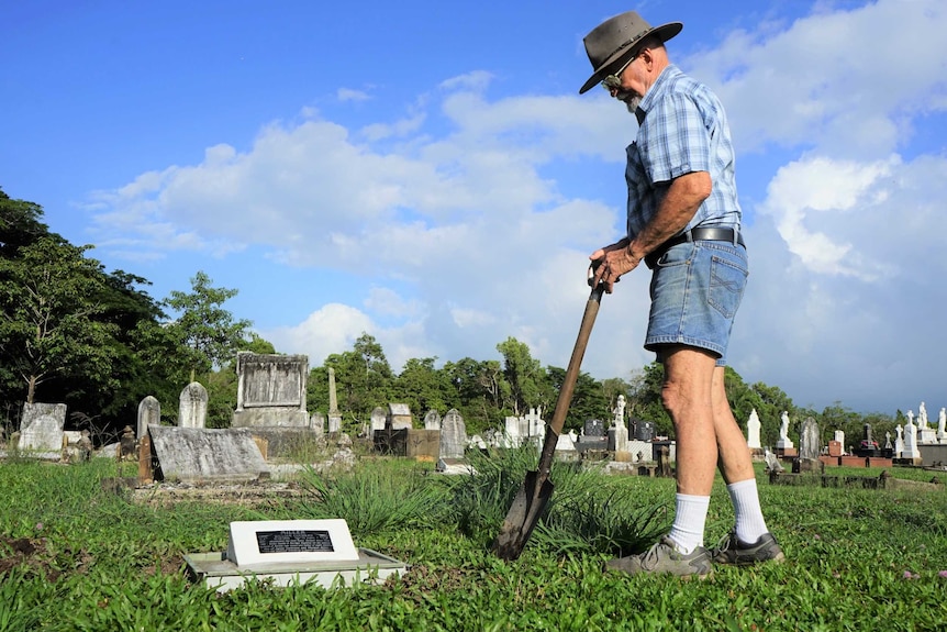 A man with a shovel stares down at a plaque in a cemetery.