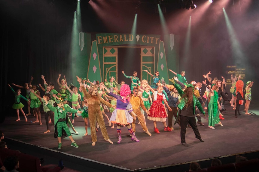 a stage with children performing in front of a green entrance door that says 'emerald city' above it.