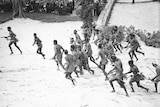 Black and white image of a group of aboriginal men wearing tradition dress, running on a beach.