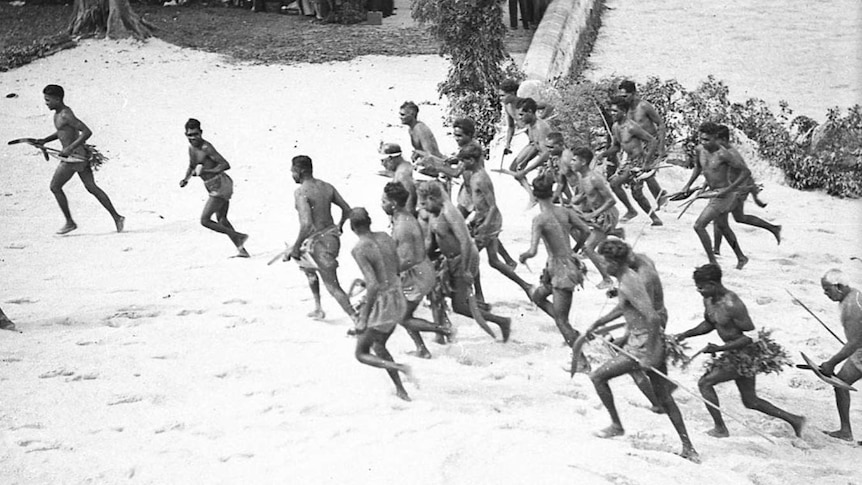 Black and white image of a group of aboriginal men wearing tradition dress, running on a beach.