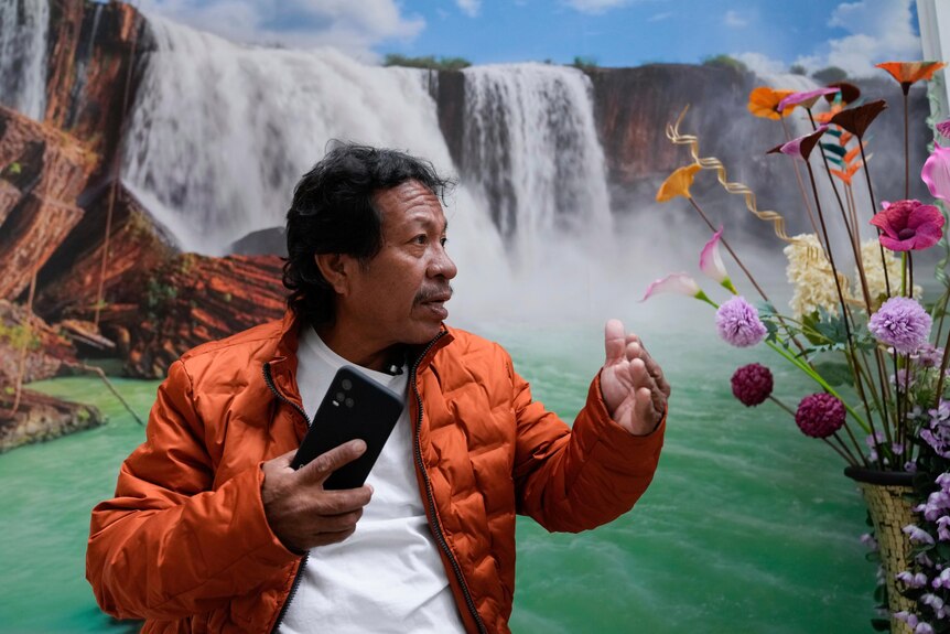 A man gestures while holding phone in front of image of waterfall. 