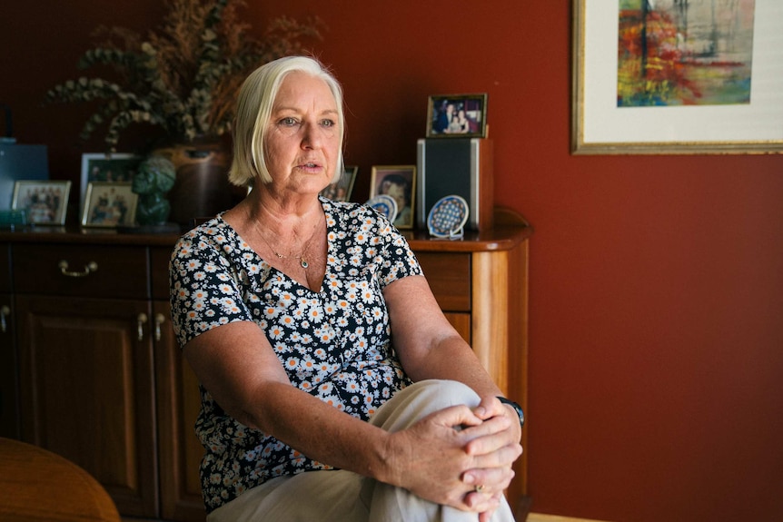 Yvonne wears a black and white floral shirt, sitting on a chair in a living room with her legs crossed and hands on her knees.