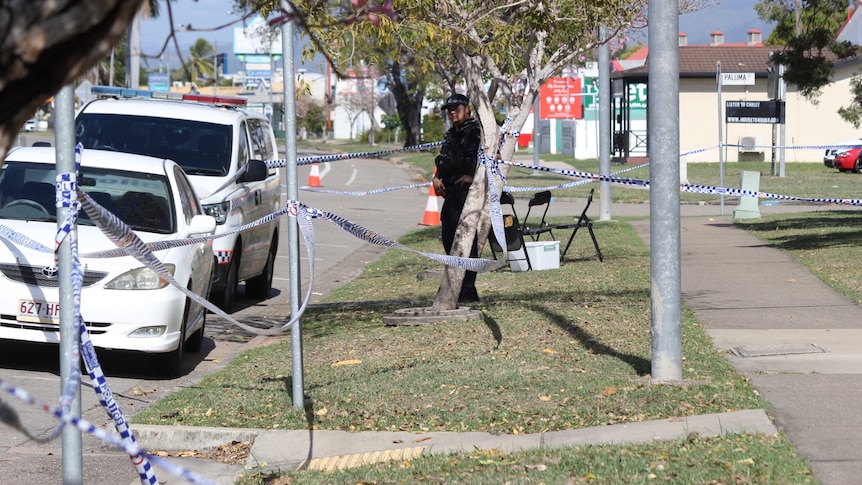 A police officer stands outside the station, with crime scene tape around nearby trees and poles.