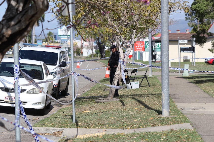 A police officer stands outside the station, with crime scene tape around nearby trees and poles.