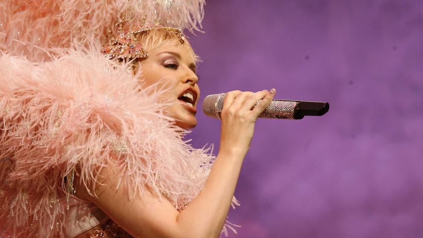 Kylie Minogue singing into a microphone in a pink, feathery burlesque costume.