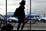 一个女人在繁忙的机场拉着一个小行李箱的剪影，通过她身后的窗户可以看到两架飞机。