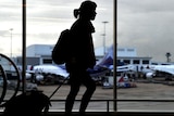 The dark silhouette of a woman pulling a small suitcase at a busy airport with two planes visible through the window behind her.