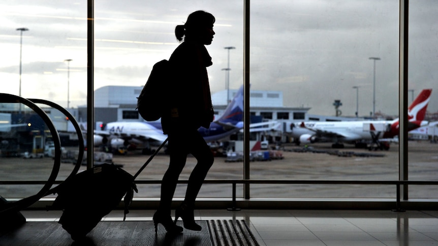 The dark silhouette of a woman pulling a small suitcase at a busy airport with two planes visible through the window behind her.