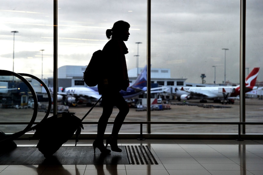 Die dunkle Silhouette einer Frau, die einen kleinen Koffer an einem belebten Flughafen zieht, mit zwei Flugzeugen, die durch das Fenster hinter ihr sichtbar sind.