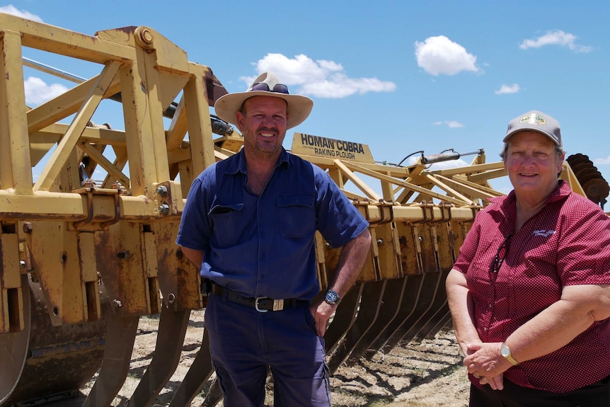 A man and woman in farming clothes stand in front of a plough