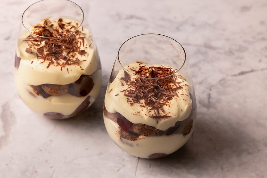 Two glasses contain layers of cream, topped with grated chocolate.
