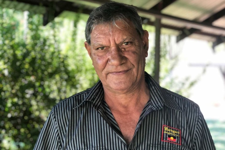 An older Indigenous man looks serious, wears a black and grey striped shirt, stands under a veranda, with green shrubs behind. 