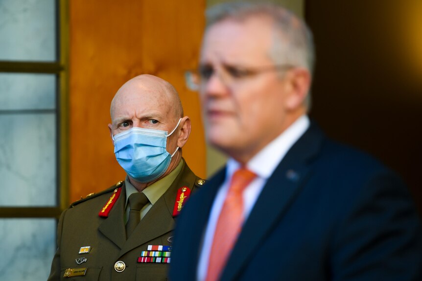 Lieutenant General John Frewen stands in the background as he looks at an out-of-focus Scott Morrison at the lectern