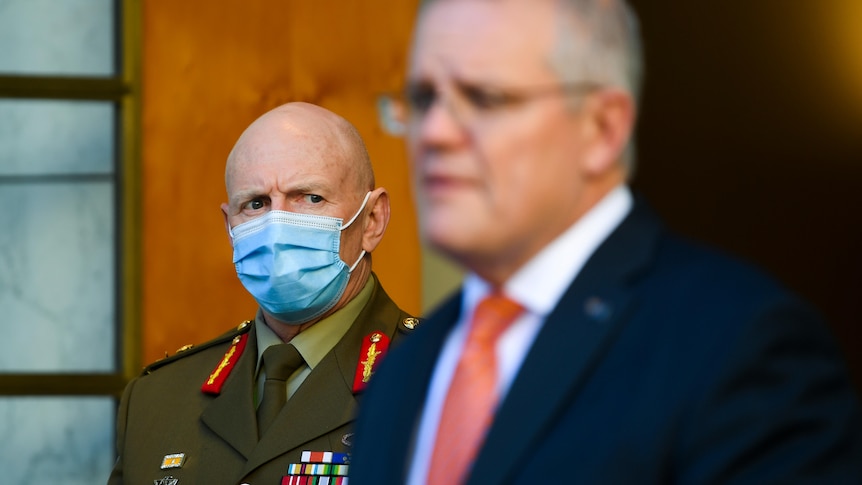 Lieutenant General John Frewen stands in the background as he looks at an out-of-focus Scott Morrison at the lectern