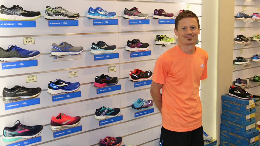 A man stands against a shop wall filled with running shoes.