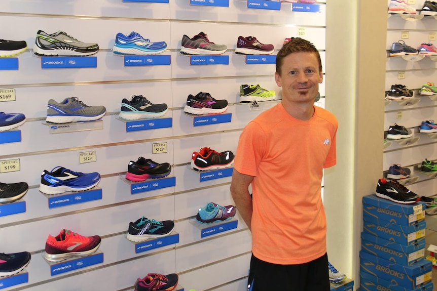 A man stands against a shop wall filled with running shoes.