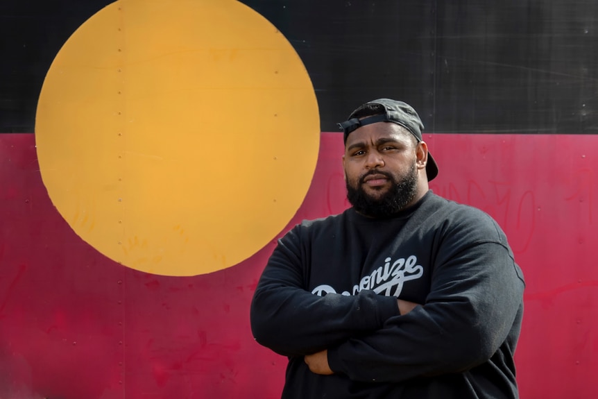 Boe Spearim crosses his arms and stands in front of an Aboriginal flag.