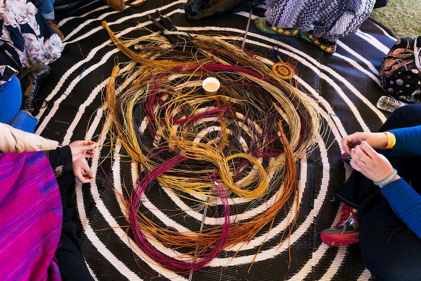 People sitting on a matt in a circle with coloured straw on the ground used to learn to weave with.