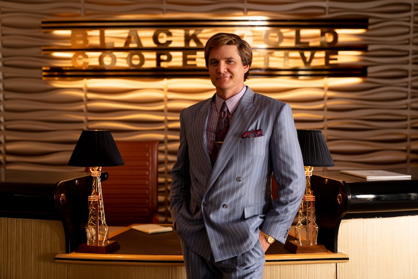 Man in grey pinstriped suit with hands in pockets, smiling suavely, with expensive looking receptionist desk behind him.