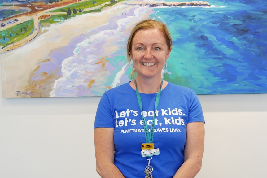 A woman wearing a blue shirt and green lanyard smiles in front of a painting of the Geraldton foreshore inside an office.