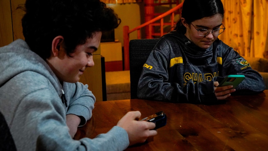 A close shot of a teenage boy and girl sitting at a timber table looking at their phones.