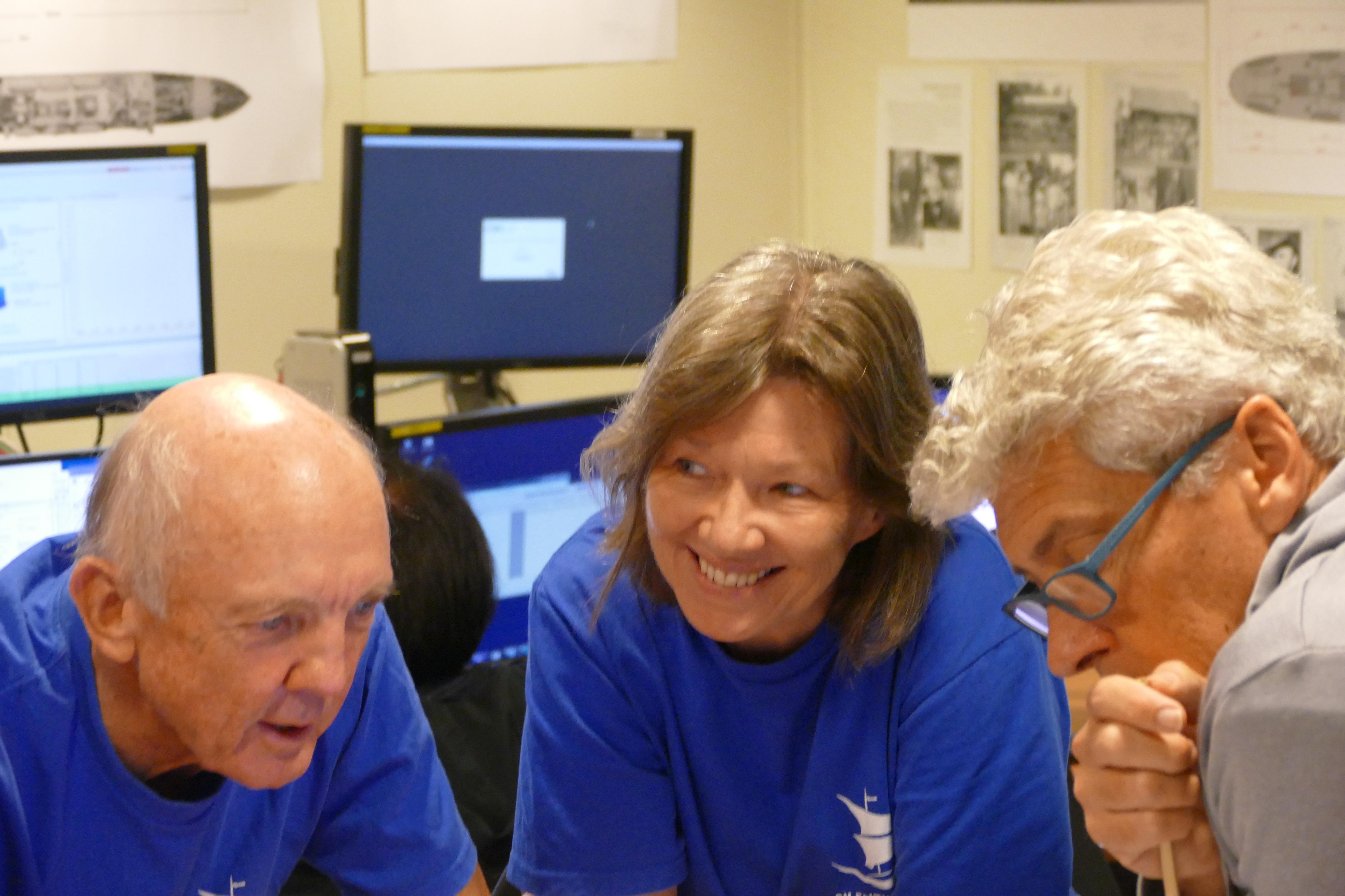 A woman smiles as she leans over a desk between two older men. 