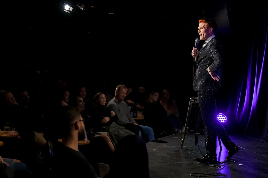 A stand-up comedian in a suit addresses a laughing audience.