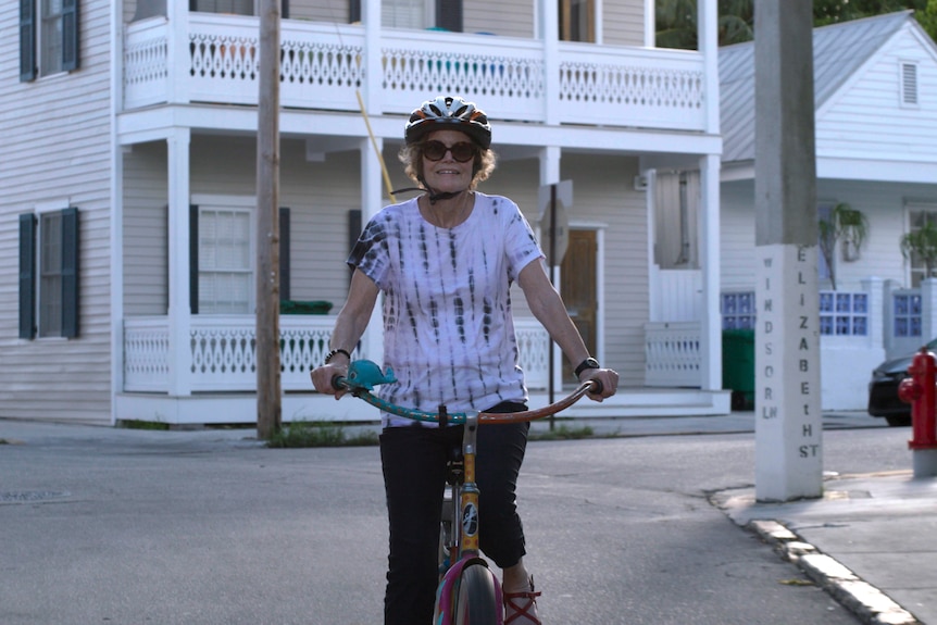 The author Judy Blume - a woman in her mid 80s in helmet and sunglasses - cycling through a town
