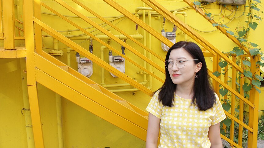 Meg O'Shea wears white and yellow checkered shirt and stands outdoors in front of yellow wall and metal stairs with some vines.