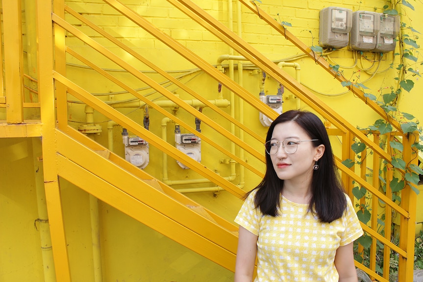 Meg O'Shea wears white and yellow checkered shirt and stands outdoors in front of yellow wall and metal stairs with some vines.