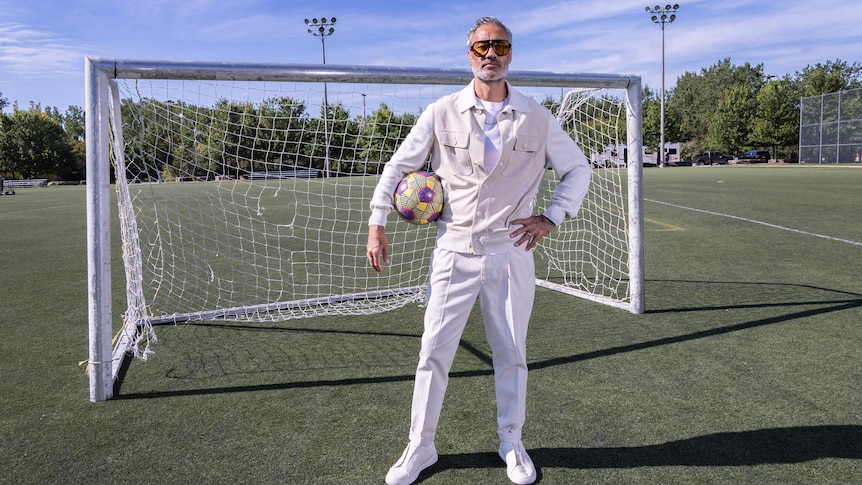 Taika Waititi stands in crisp white clothes and sunnies, in front of a soccer net on a field, ball under one arm