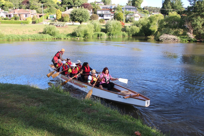 Eight people of different ages and walks of life sit in a dragon boat on a river, all smiling.