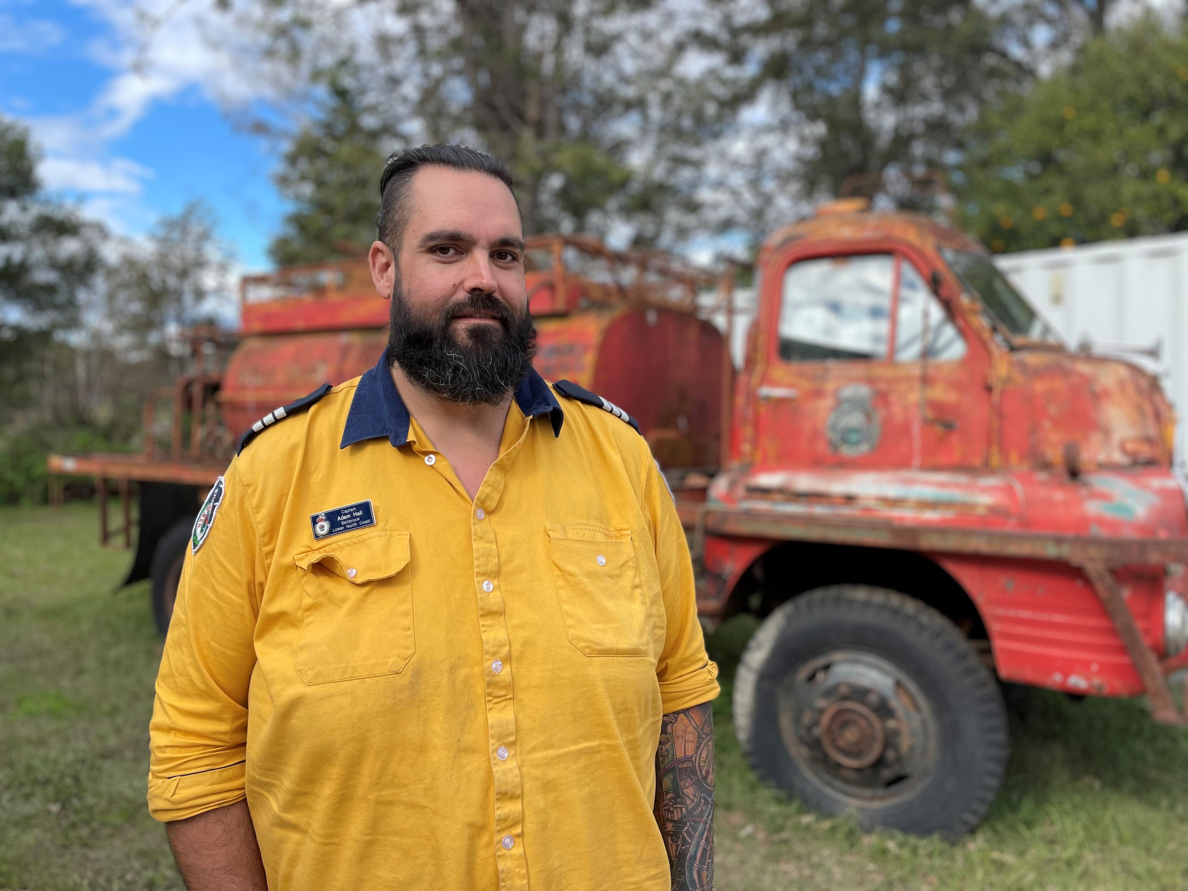 A man in a yellow firefighter shirt stands in front of an old truck.