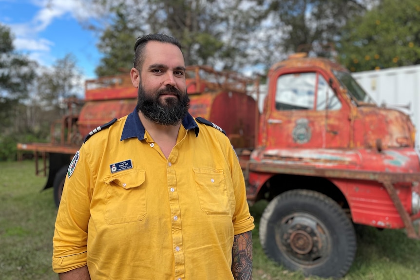 A man in a yellow firefighter shirt stands in front of an old truck.