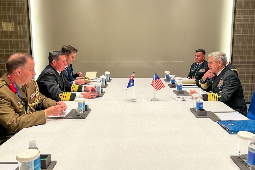 Men in military uniforms sit around a table, miniature Australian and US flags in the centre.