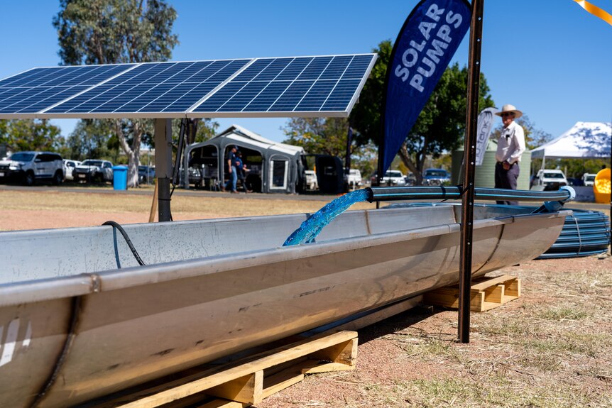 Water rushes from a pipe into a trough. In the background is a large solar panel and a flag that says "solar pumps".